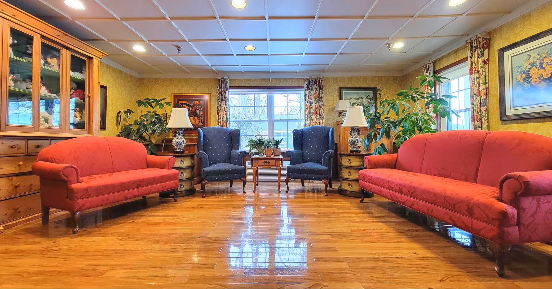 A cozy communal sitting room with two red sofas, two blue armchairs, wooden floors, plants, lamps and framed art arranged around a central window.