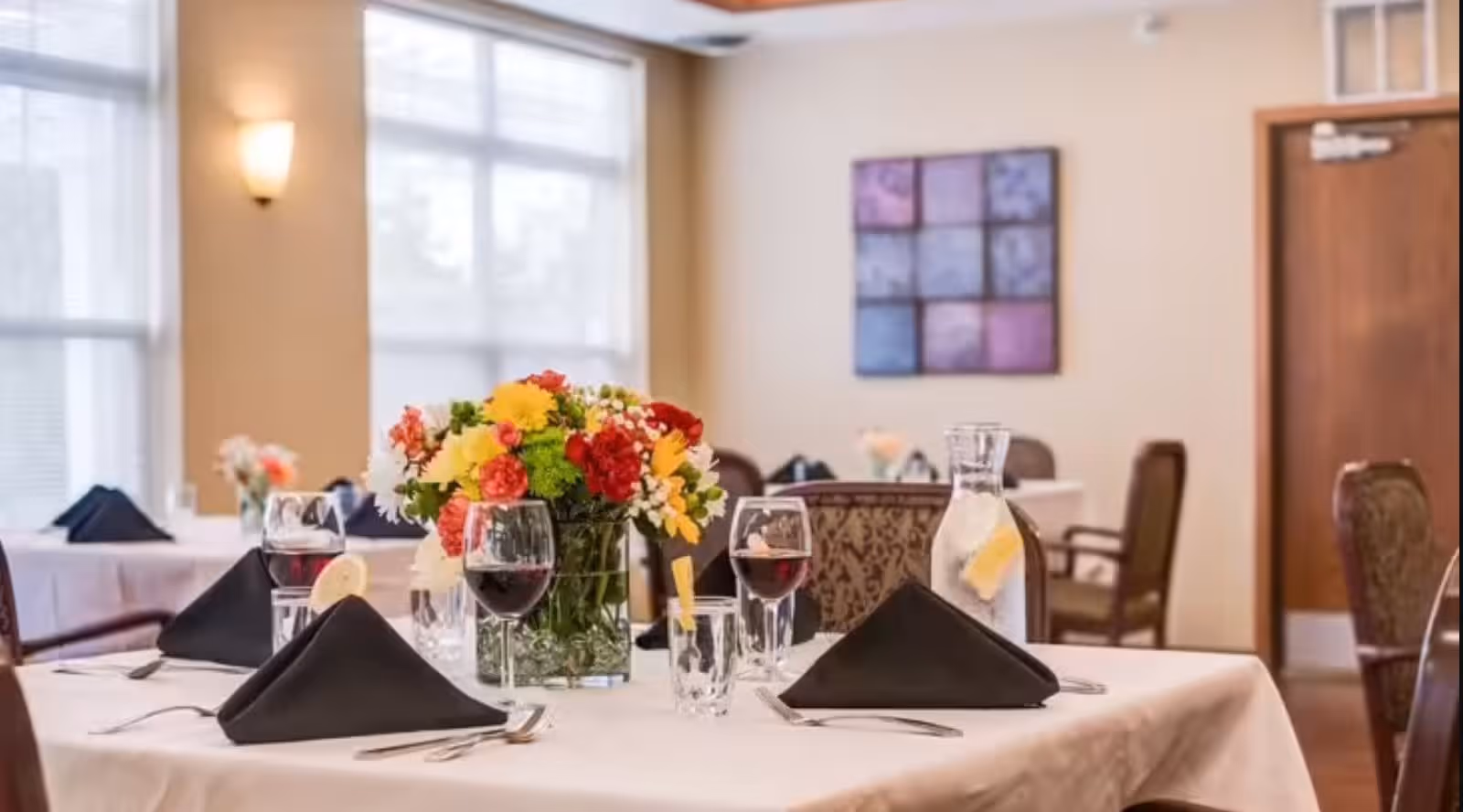 Dining room table set with a floral centerpiece, wine glasses, and black napkins in a bright communal dining area.