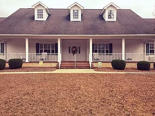 Front exterior view of a single-story house with a large porch, three dormer windows on the roof, white railings, and neatly trimmed bushes in front.