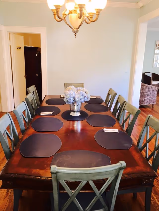 A dining room with a long wooden table set with eight black placemats and napkins. A silver vase with light purple flowers is centered on the table. The room has light blue walls, wooden flooring, and a chandelier hanging above the table. There are green wooden chairs around the table, and an open doorway leads to another room with wicker furniture.