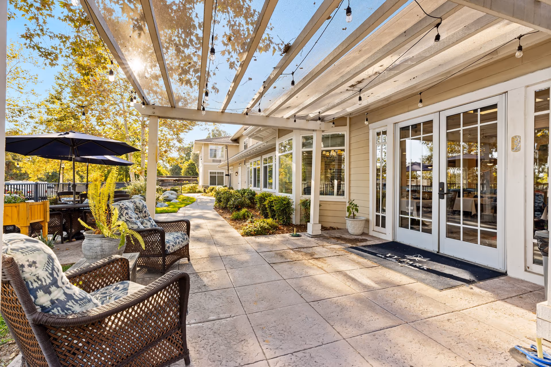 Outdoor patio area at Ivy Park at Alta Loma with wicker chairs featuring patterned cushions, potted plants, a pergola with string lights, and a pathway leading to the building entrance with glass double doors.