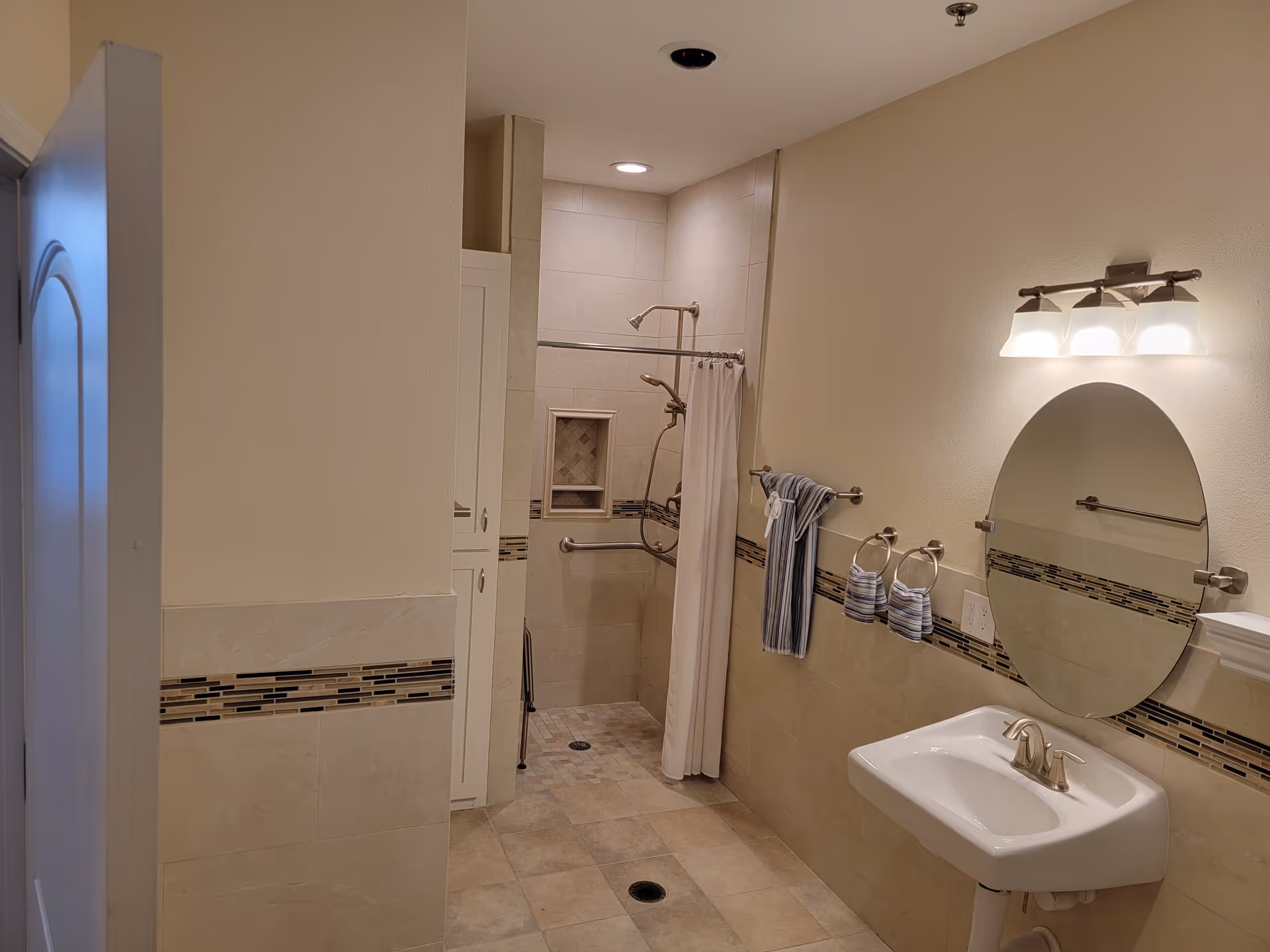 Interior view of a bathroom with beige tiled walls and floor. The bathroom features a walk-in shower with a curtain, a built-in niche, and grab bars. There is a white pedestal sink with a round mirror above it and a three-light fixture. Towels hang on a towel bar and rings next to the sink.
