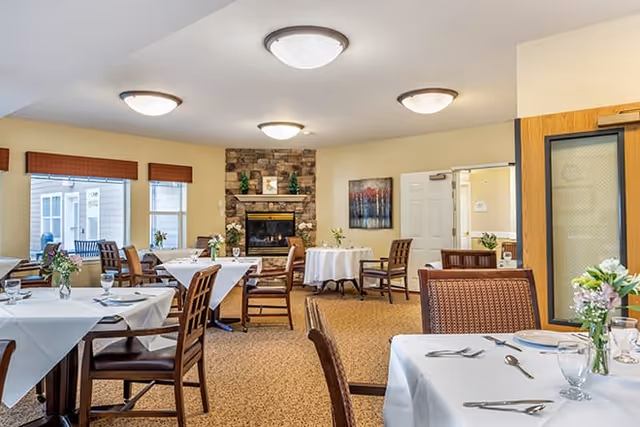 A bright dining room with several tables covered in white tablecloths, each set with plates, silverware, glasses, and small floral centerpieces. The room features a stone fireplace, multiple windows with brown blinds, and warm lighting from ceiling fixtures. Wooden chairs are arranged around the tables, and there is a painting on the wall near a door.