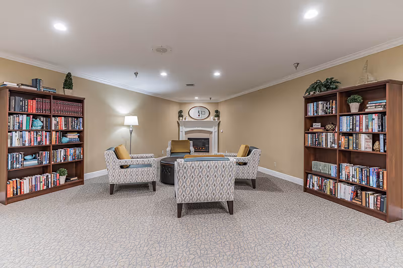 A cozy reading and sitting area with four patterned armchairs arranged around a round black ottoman. There are two wooden bookshelves filled with books on either side of the room. A floor lamp stands near the left bookshelf, and a fireplace with decorative items on the mantel is centered on the back wall. The room has beige walls and carpeted flooring.