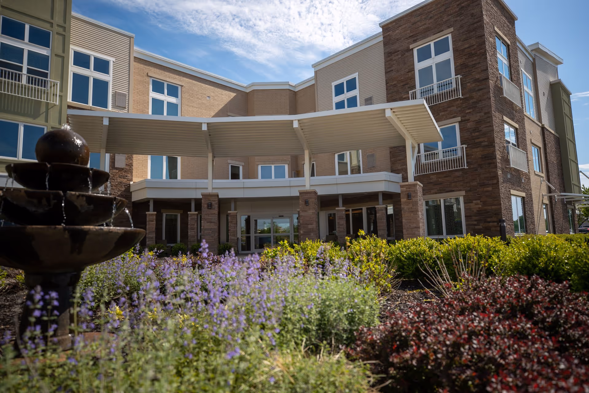 Exterior view of Vista Springs Greenbriar Village building with a covered entrance, large windows, and a landscaped garden featuring purple flowers and a tiered water fountain in the foreground under a partly cloudy sky.