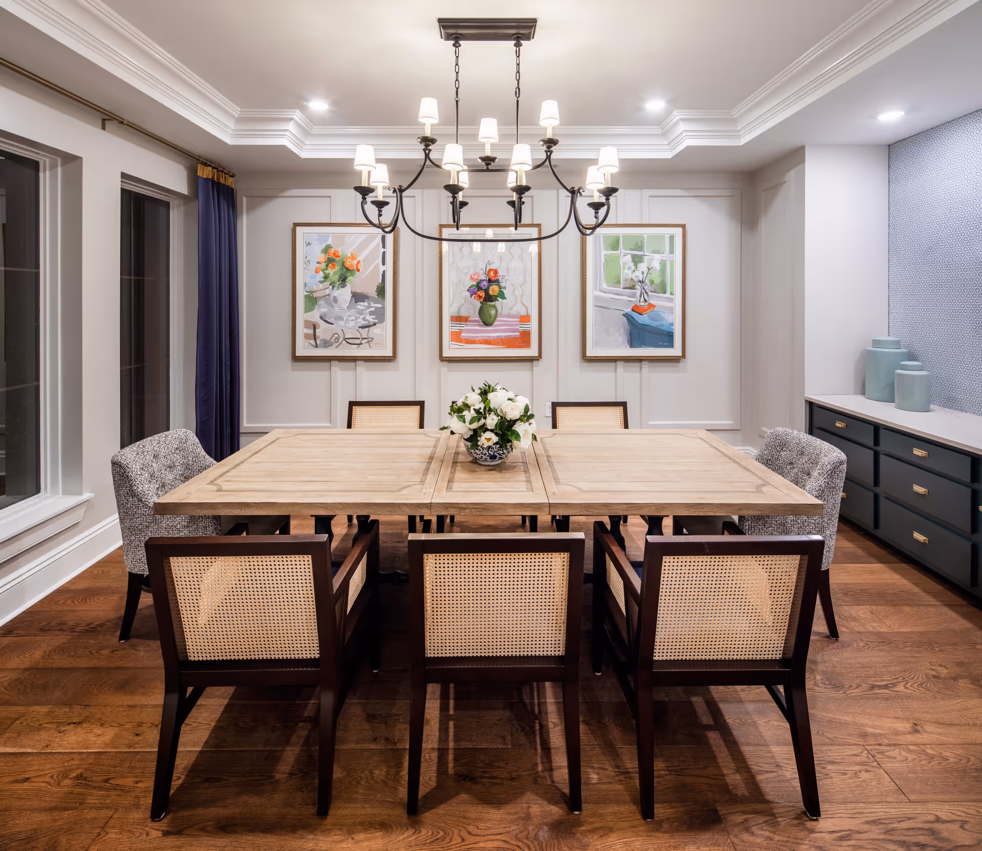 A well-lit dining room with a large wooden table surrounded by six chairs, three with cane backs and three upholstered. A chandelier with multiple lights hangs above the table. The walls are decorated with three framed paintings of floral arrangements. There are large windows with dark curtains on the left side and a sideboard with decorative vases on the right.