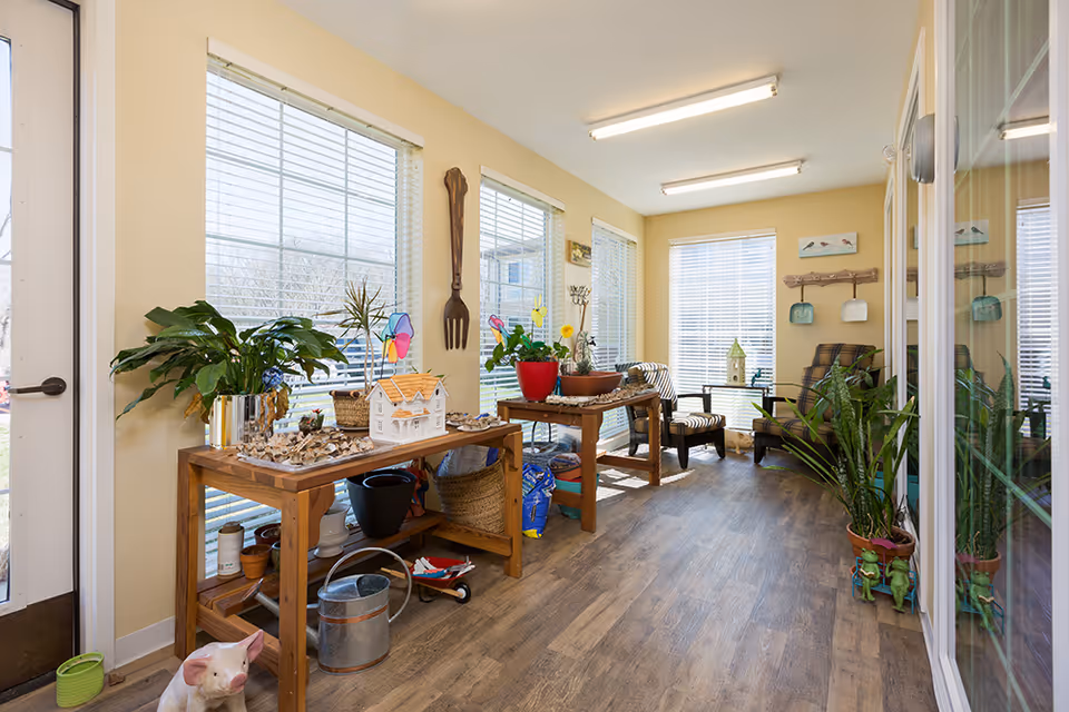 Bright sunroom-style common sitting area with wooden tables, potted plants, chairs and large windows letting in natural light.
