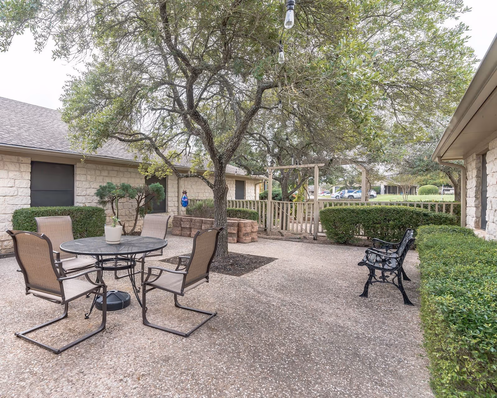 Outdoor patio area with a round metal table and four chairs, a large tree in the center, two black metal benches on the right, surrounded by trimmed bushes and stone buildings in the background.