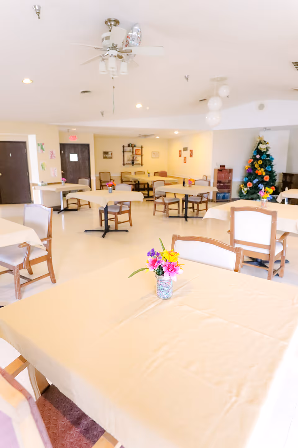 A bright communal dining room with tables covered in beige tablecloths and small flower centerpieces and a decorated Christmas tree in the background.