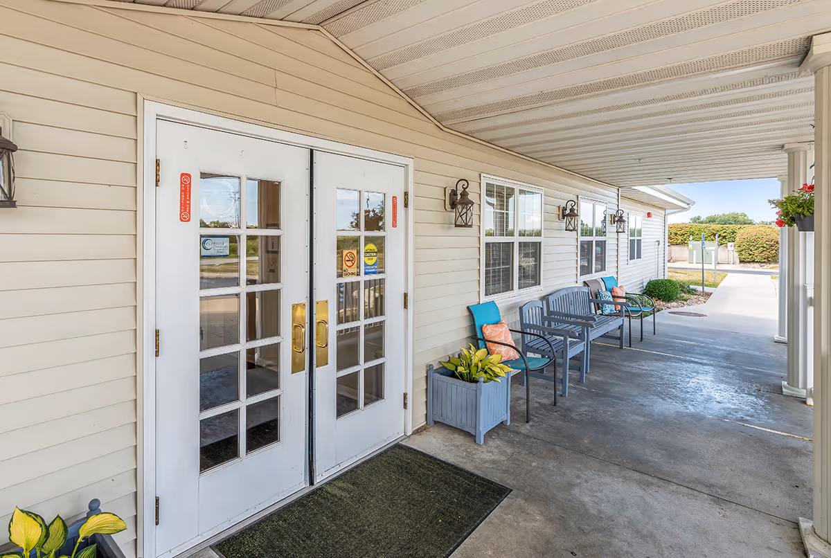 Covered entrance area of Eiler Senior Living with white double doors featuring glass panes and brass handles. Along the right side, there are several gray benches and chairs with colorful cushions, potted plants, and wall-mounted lantern-style lights. The area opens to a paved walkway and greenery in the background.