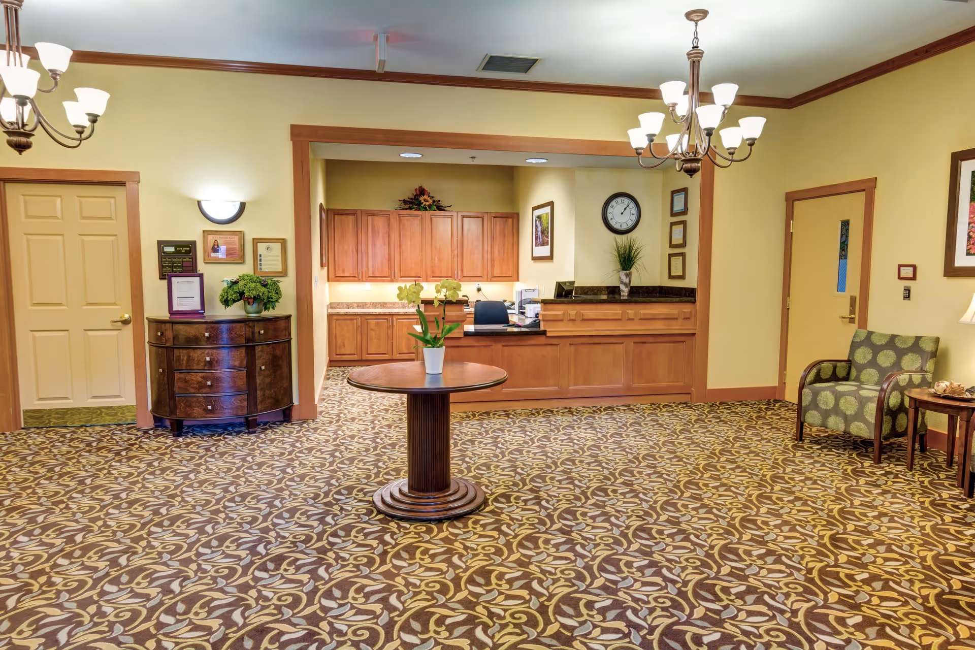 Reception area of Rosewood Memory Care facility with a wooden front desk, a round table with a potted plant in the center, patterned carpet, a green armchair with a side table and lamp, wall clock, framed pictures, and warm yellow walls with wooden trim.