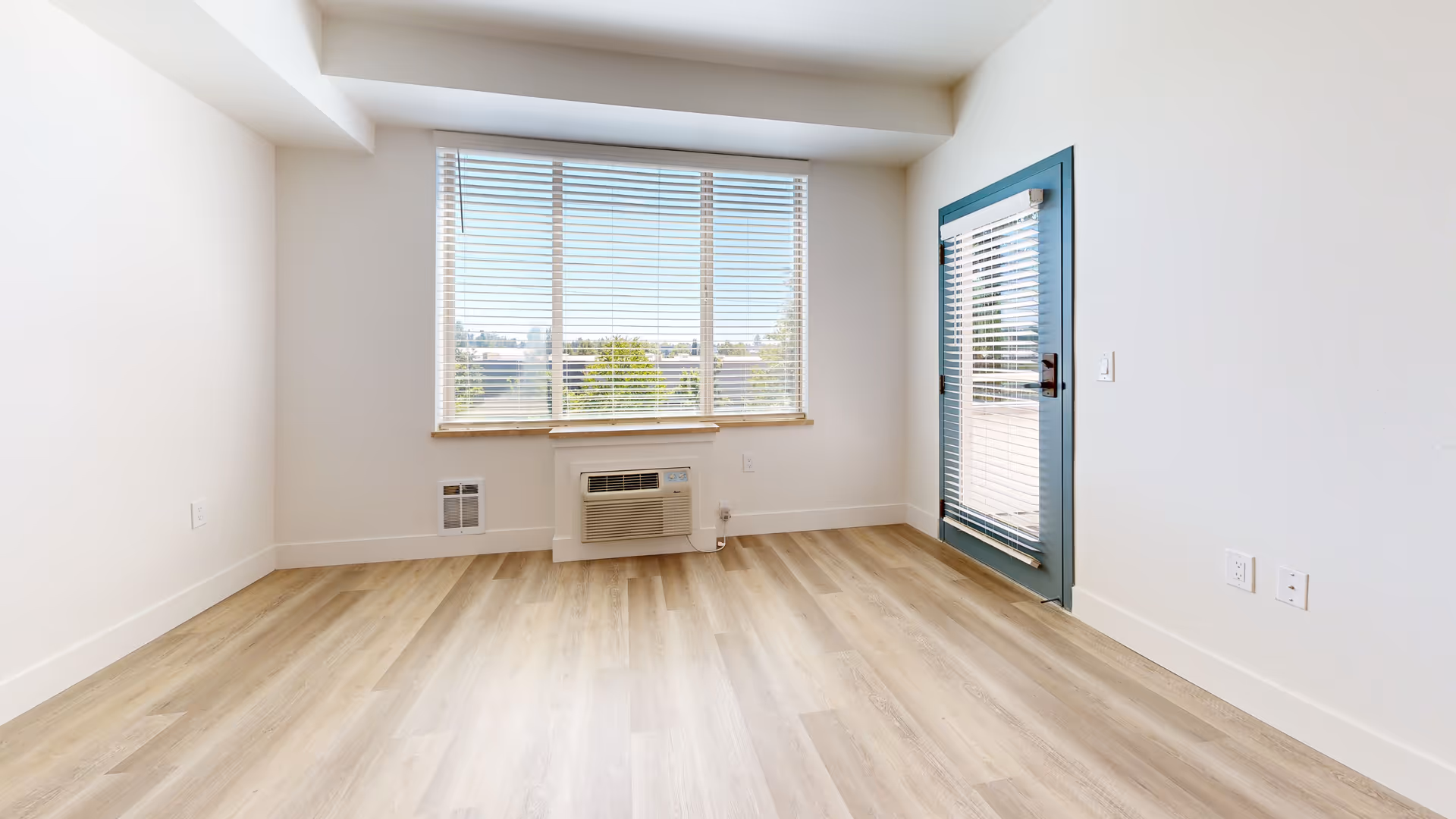 Empty room with light wood flooring, white walls, a large window with blinds, and a blue-framed glass door with blinds leading outside.