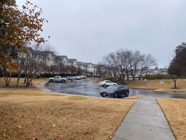 View of a senior living facility parking lot on a cloudy day with several parked cars, leafless trees, and a large multi-story building in the background. The foreground shows a concrete sidewalk and brown grass.
