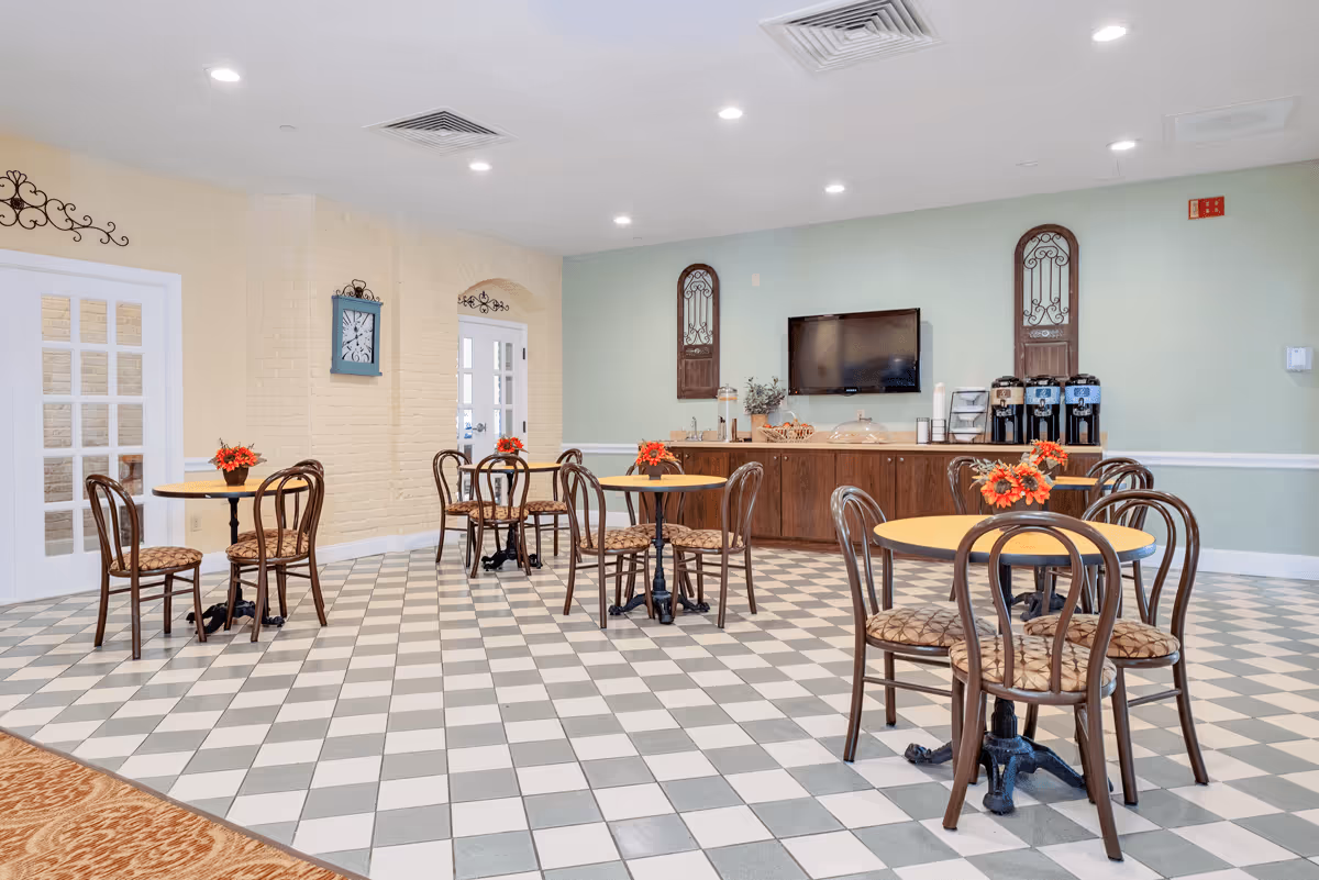 Bright communal dining area with round tables and chairs on a checkered floor, a TV and coffee station along the back wall, and small floral centerpieces on each table.