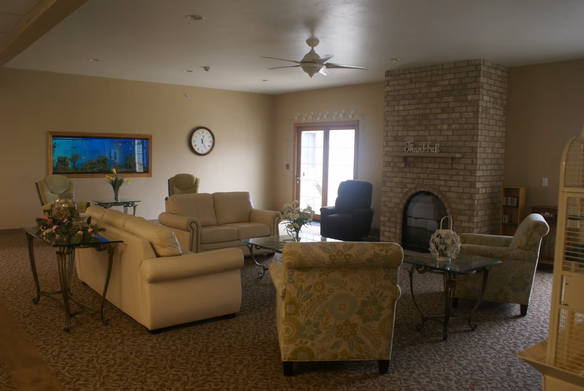 A cozy living room with beige sofas and floral armchairs arranged around glass coffee tables. There is a brick fireplace with the word 'Thankful' mounted above it, a wall clock, a large aquarium, and a ceiling fan. French doors let in natural light.