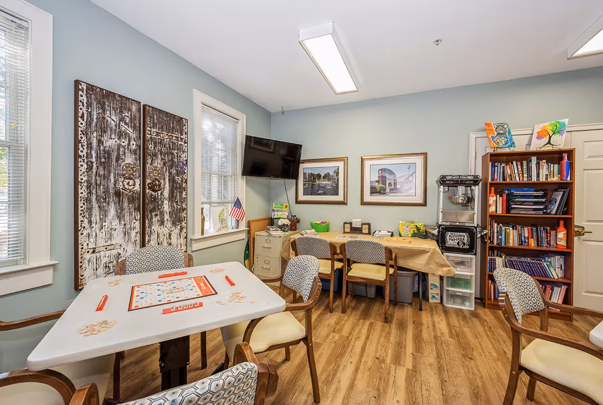 A cozy activity room with a table set up for a game of Scrabble, surrounded by four cushioned chairs. The room has light blue walls, two windows with blinds, and two large rustic wooden panels on one wall. A flat-screen TV is mounted in the corner above a filing cabinet. A table covered with a beige cloth holds various items including a popcorn machine and framed pictures on the wall above it. A wooden bookshelf filled with books and colorful artwork on top is positioned near a closed door. The floor is wood laminate.