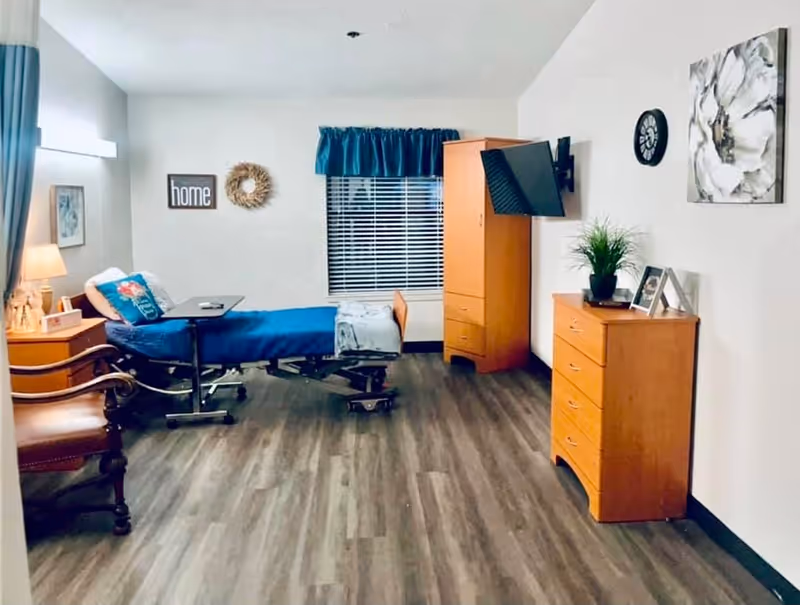 Furnished senior care bedroom with a hospital-style bed, wooden dressers, a wall-mounted TV, chair, and window with blinds.