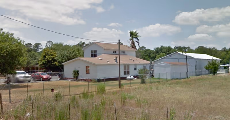 Exterior view of a two-story white building with a brown roof, surrounded by a grassy field and a chain-link fence. There are several parked cars and trees in the background under a partly cloudy sky.