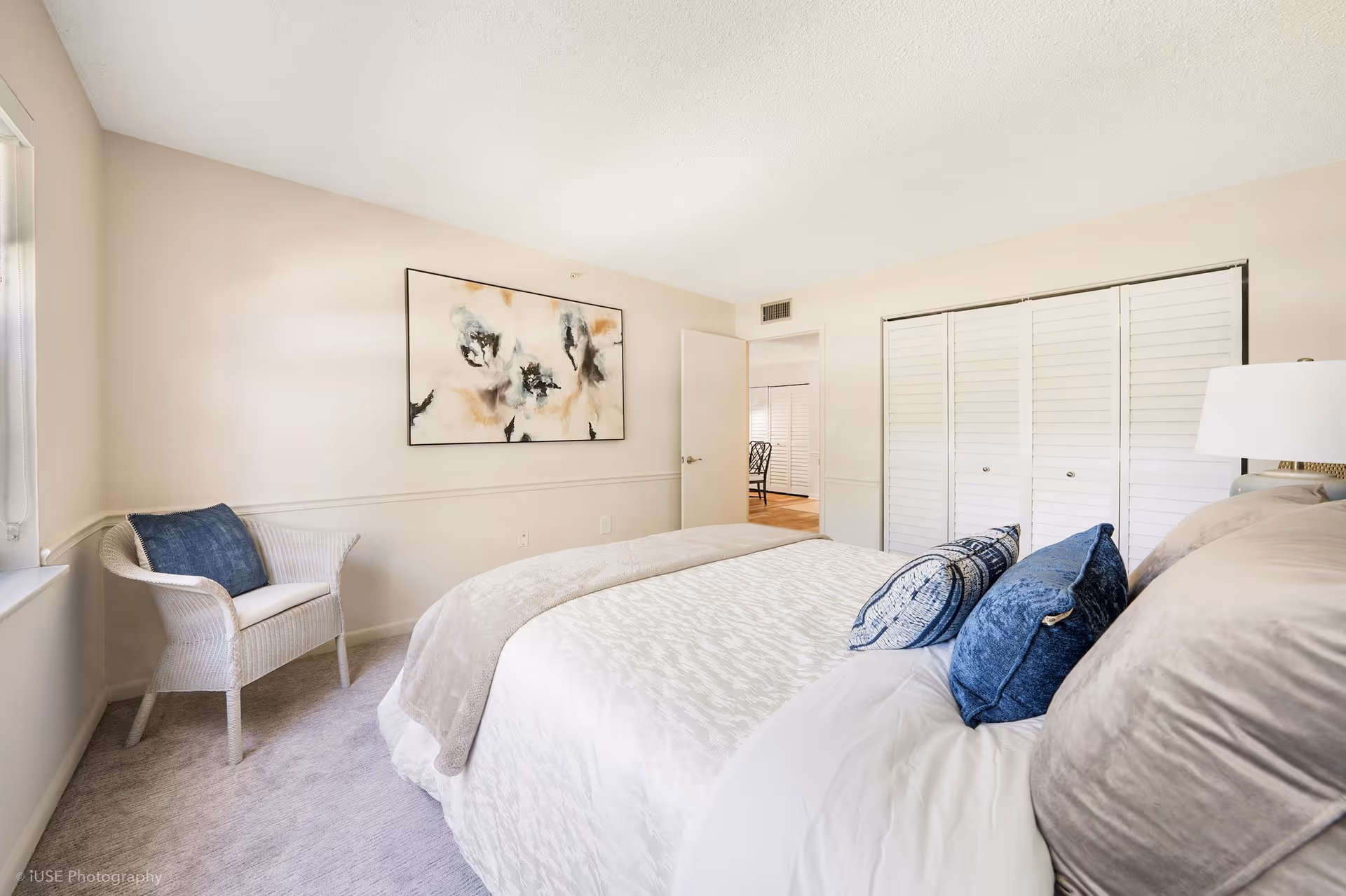 A bright and cozy bedroom with a large bed adorned with beige and blue pillows and a beige blanket. There is a white wicker chair with a blue cushion near a window on the left. A large abstract painting hangs on the wall above the chair. The room has light-colored walls, a carpeted floor, and white louvered closet doors. The door to the room is open, showing a glimpse of another room with wooden flooring and a chair.