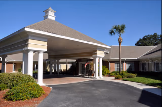 Entrance of a senior living facility with a covered driveway supported by white columns, surrounded by landscaped bushes and a palm tree under a clear blue sky.