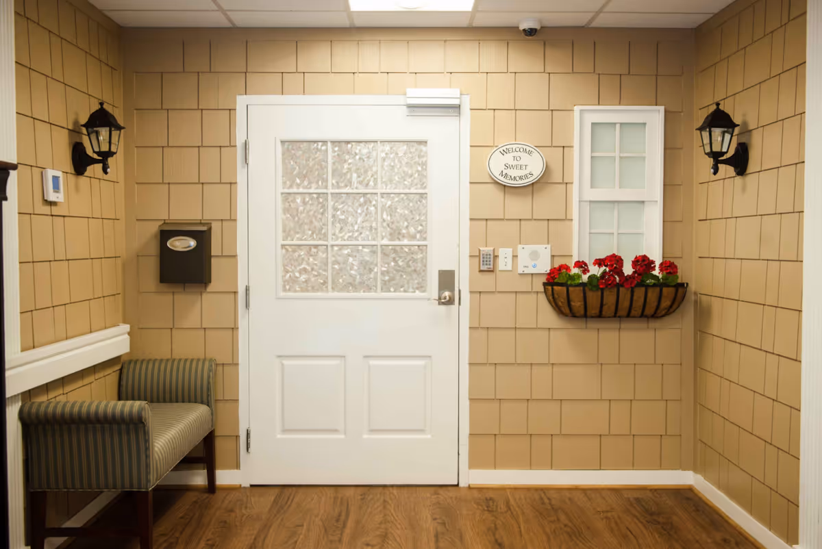 Interior view of a hallway or entry area with beige shingle walls, a white door with frosted glass panels, a small window with frosted glass, a flower box with red flowers, two black wall-mounted lantern-style lights, a striped upholstered bench, a mailbox labeled 'Mail', and a sign that reads 'Welcome to Sweet Memories'.