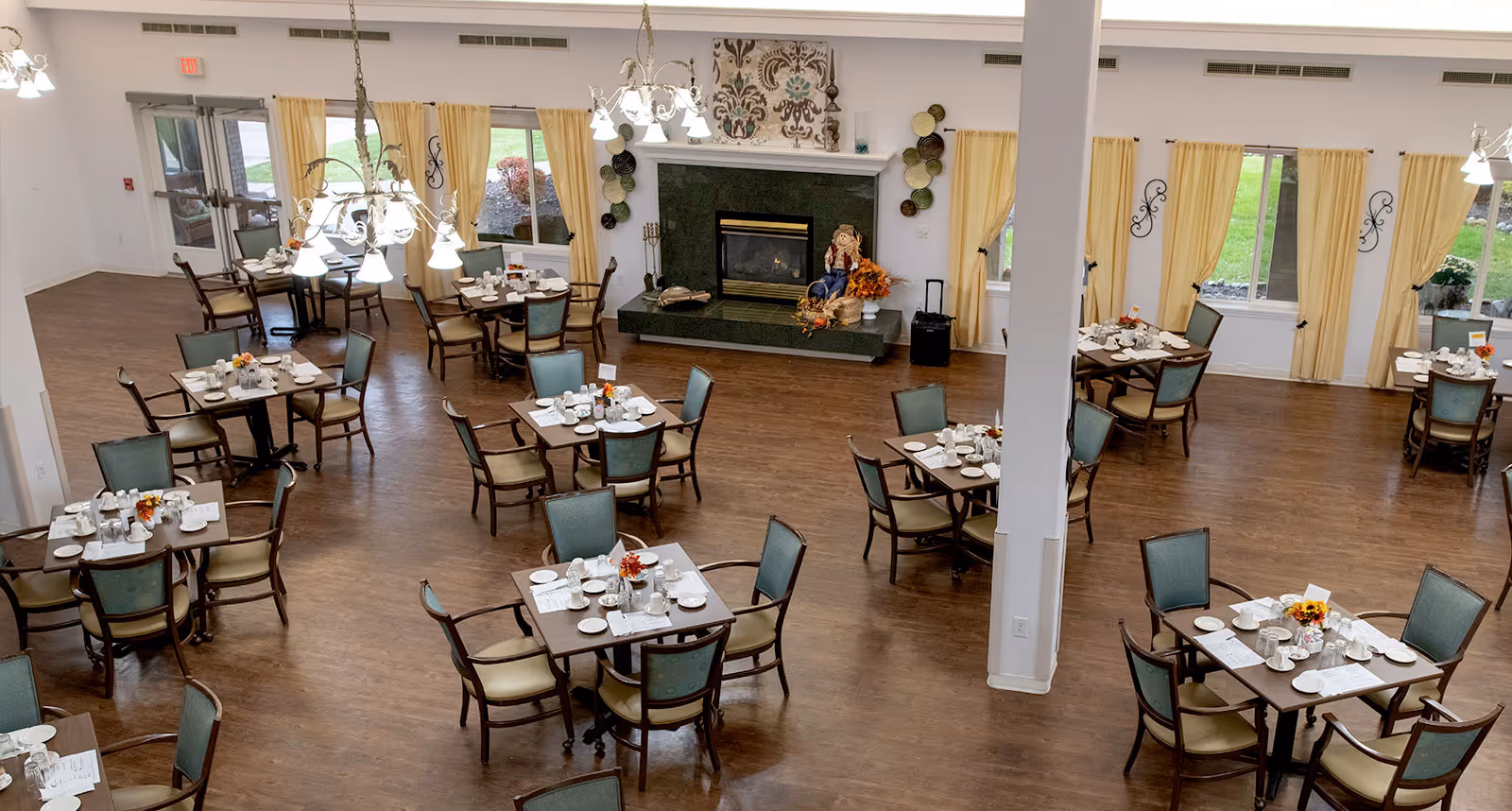 A spacious dining room with multiple square tables set for meals, each table surrounded by four chairs. The room features wooden flooring, large windows with yellow curtains, and a fireplace decorated with a scarecrow and autumn-themed items. Chandeliers hang from the ceiling providing warm lighting.