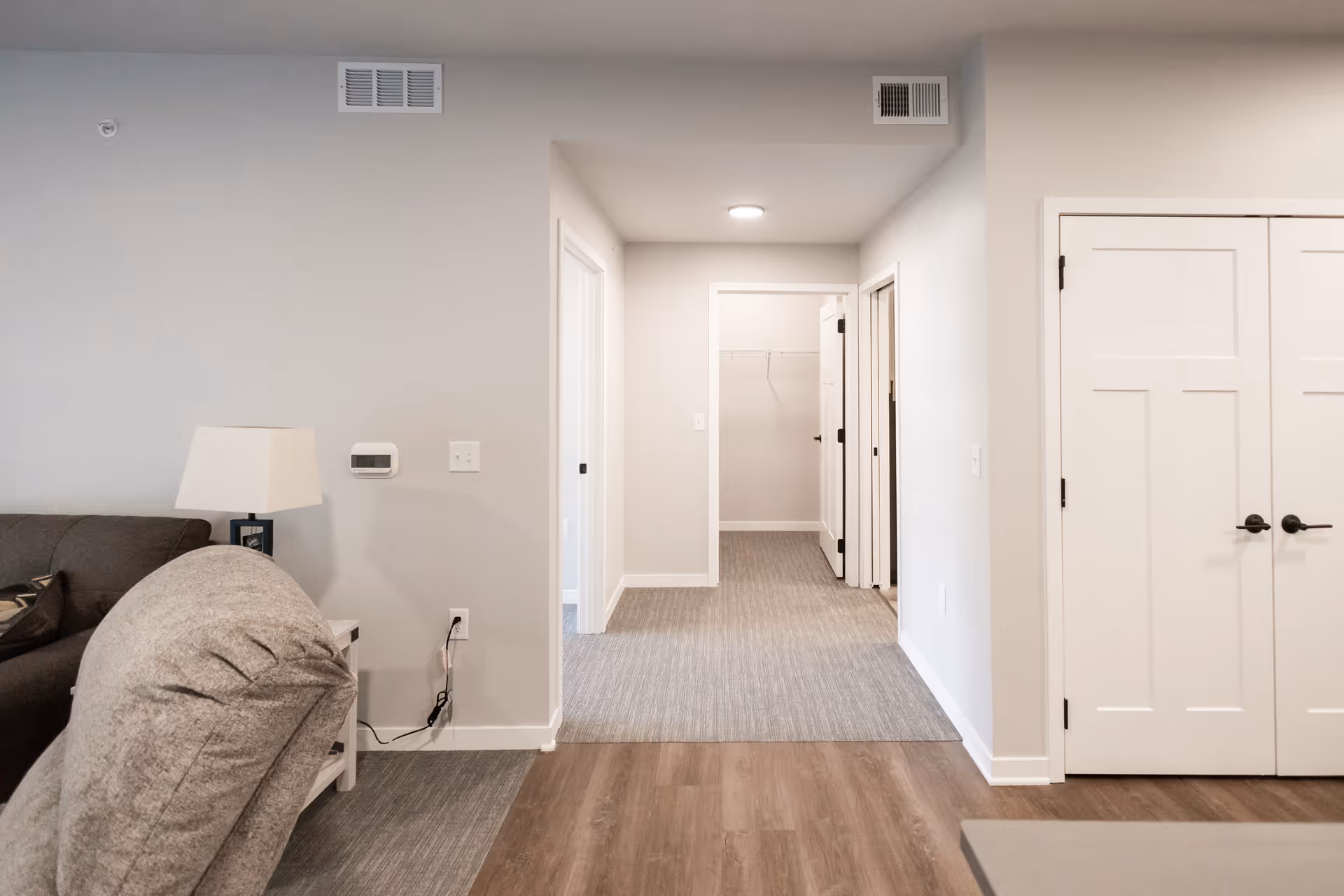 Interior view of a senior living facility hallway with light-colored walls and carpeted floor. There is a gray armchair and a side table with a lamp on the left side. The hallway leads to an open closet with a wire shelf and two closed doors on the right. The floor transitions from wood to carpet near the hallway.