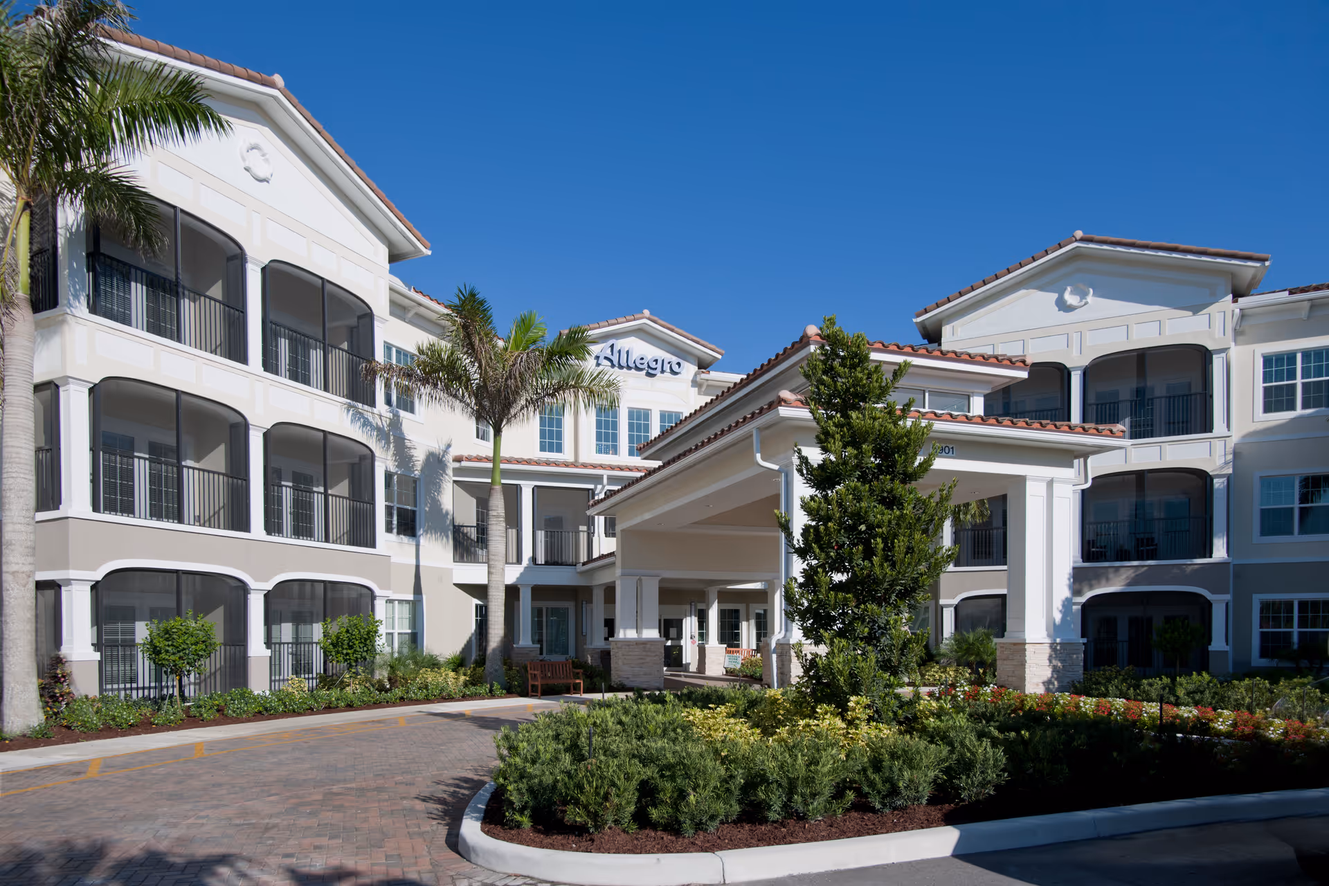 Exterior view of Allegro Senior Living facility in Parkland, FL, showing a multi-story building with balconies, palm trees, landscaped greenery, and a covered entrance under a clear blue sky.