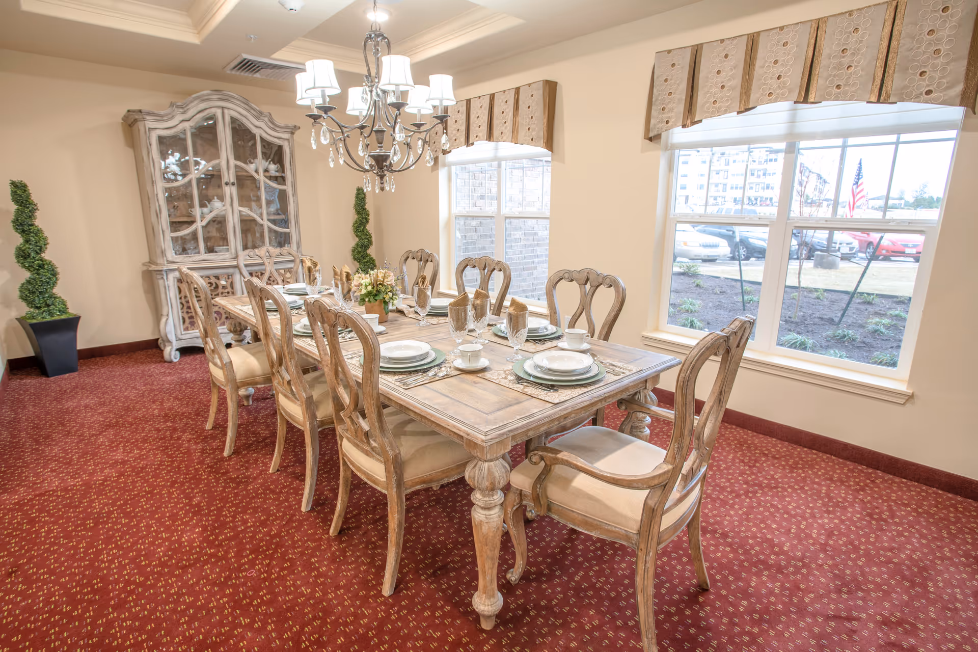 A formal dining room with a wooden dining table set for eight people. The table is adorned with plates, cups, glasses, and folded napkins. There is a chandelier hanging above the table, two windows with valances letting in natural light, a decorative cabinet in the corner, and two spiral-shaped topiary plants in black pots. The room has beige walls and a red patterned carpet.