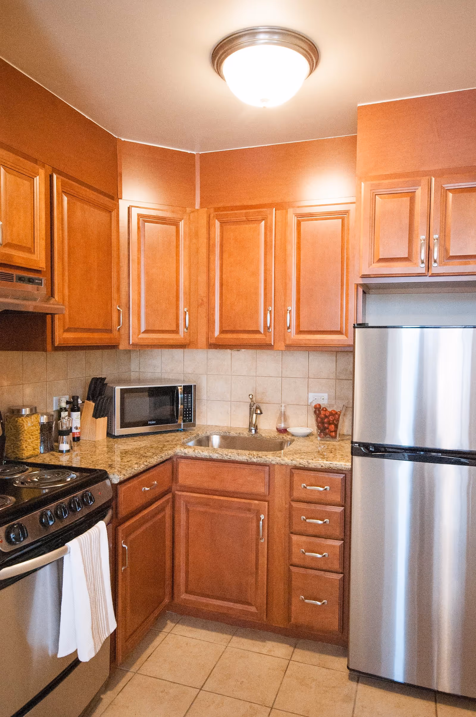 A compact kitchen with wooden cabinets, a granite countertop, a stainless steel refrigerator, a microwave, a stove with a white towel hanging on the handle, a sink, and various kitchen items including a knife block and jars on the counter.