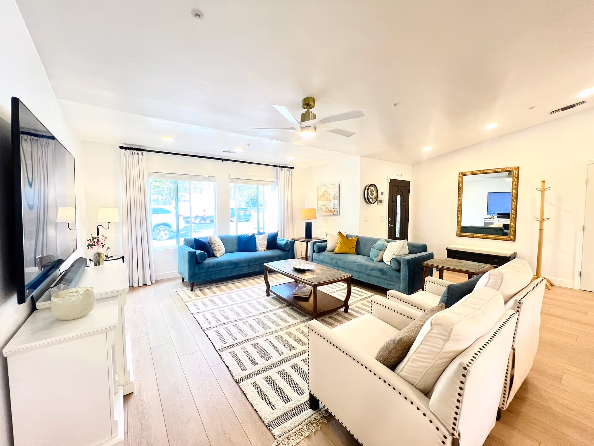 Bright and spacious living room with two teal sofas, two white armchairs, a wooden coffee table on a patterned rug, a large flat-screen TV mounted on the wall, a ceiling fan, and large windows with white curtains letting in natural light.