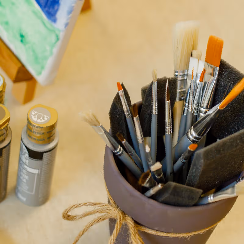 Close-up view of a container holding various paintbrushes of different sizes and shapes, with paint bottles and a partially visible canvas in the background.