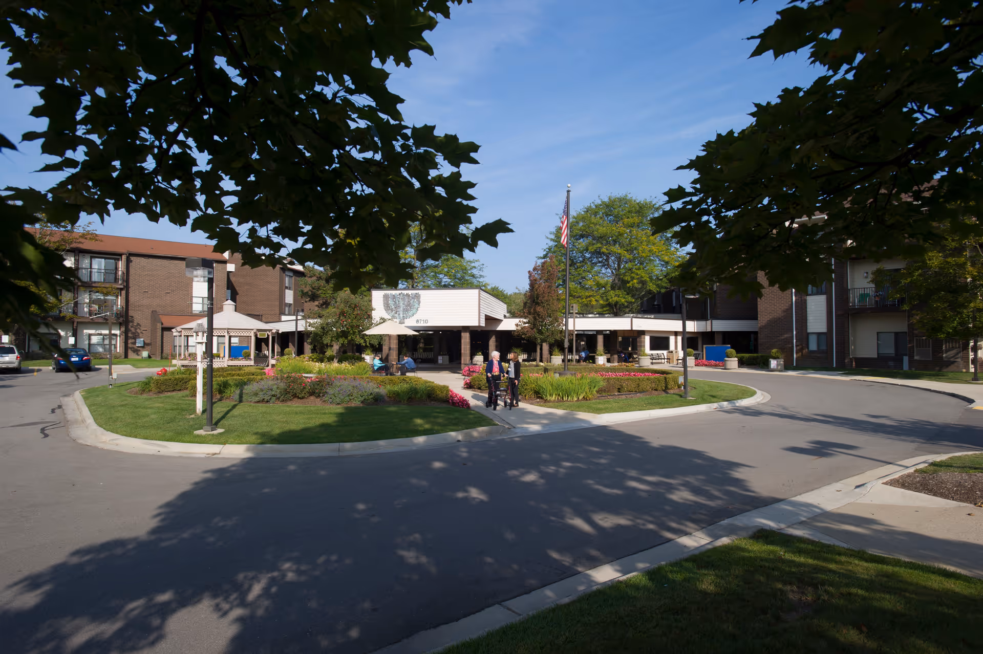 Exterior view of the Jewish Senior Life facility showing a circular driveway with landscaped greenery and flowers in the center. The building is a multi-story brick structure with balconies and an entrance canopy. Two people are walking on the pathway towards the entrance. Trees frame the image with a clear blue sky above.
