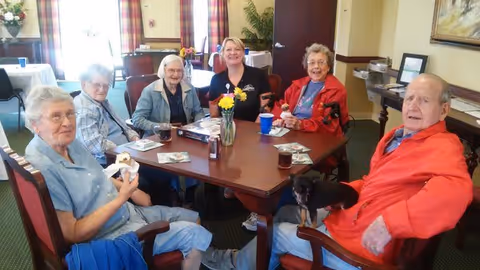 A group of five elderly individuals and one staff member sitting around a wooden table in a dining area. The table has a vase with yellow flowers, drinks, and some papers. The room has large windows with plaid curtains and other tables and chairs in the background.