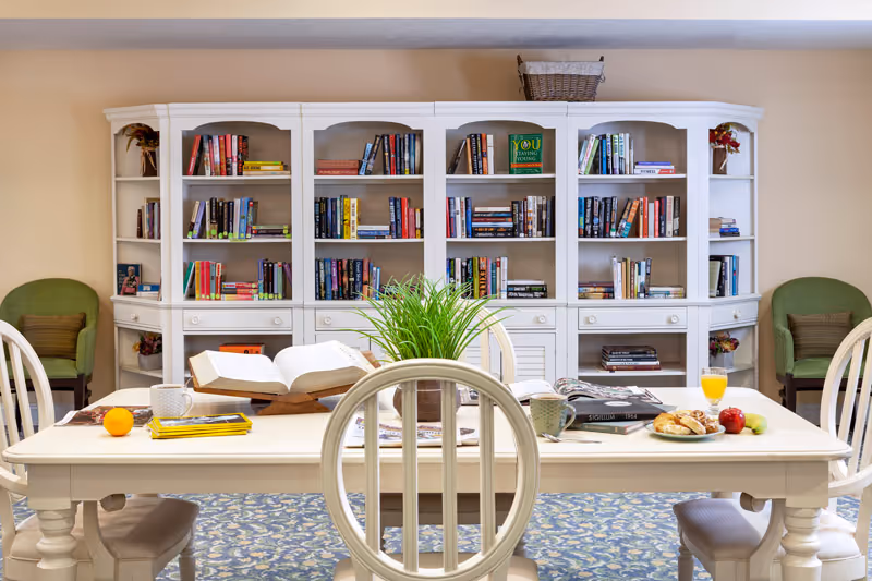 A cozy reading and dining area with a white wooden table surrounded by chairs. On the table are books, a plant, a cup, a glass of orange juice, and a plate with pastries and fruit. Behind the table is a large white bookshelf filled with books and decorative items. Two green cushioned chairs are placed against the beige wall on either side of the bookshelf.