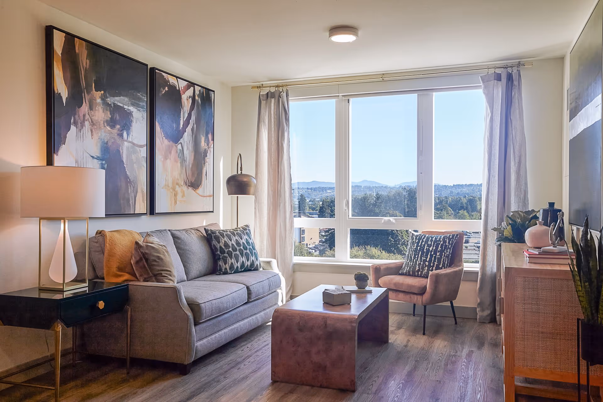A bright living room with a large window showing a scenic view of trees and distant hills. The room features a gray sofa with patterned and solid pillows, a modern floor lamp, two abstract paintings on the wall, a brown armchair with a patterned pillow, a wooden coffee table with decorative items, and a wooden cabinet with plants and books on top. Light curtains frame the window and the floor is wooden.