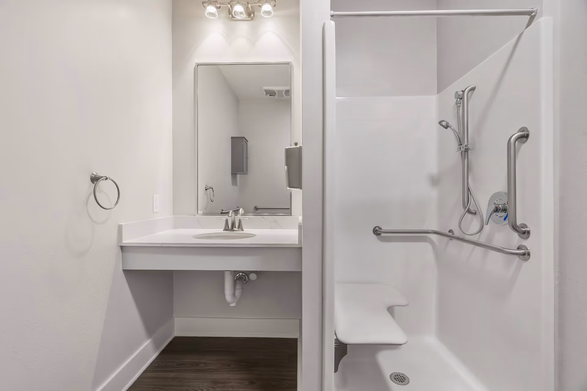 Accessible bathroom featuring a sink and mirror on the left and a shower with grab bars and a fold-down seat on the right.