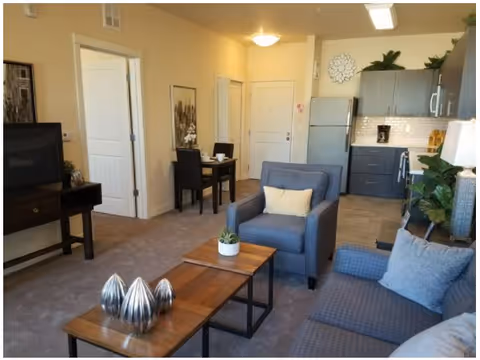 Interior view of a senior living facility apartment featuring a living area with a blue armchair and sofa, a wooden coffee table with decorative items, a small dining table with two chairs, and a kitchen area with gray cabinets, a refrigerator, and a microwave. The walls are light-colored, and there are decorative plants and artwork on the walls.