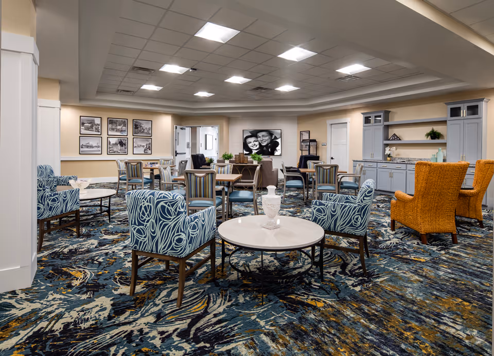 A spacious and well-lit common area in a senior living facility with multiple seating arrangements including blue patterned armchairs, wicker chairs, and tables. The room features a colorful carpet with abstract patterns, light-colored walls, framed black and white photos on one wall, and a large black and white portrait on another. There is cabinetry with decorative items and plants, and a ceiling with recessed lighting.