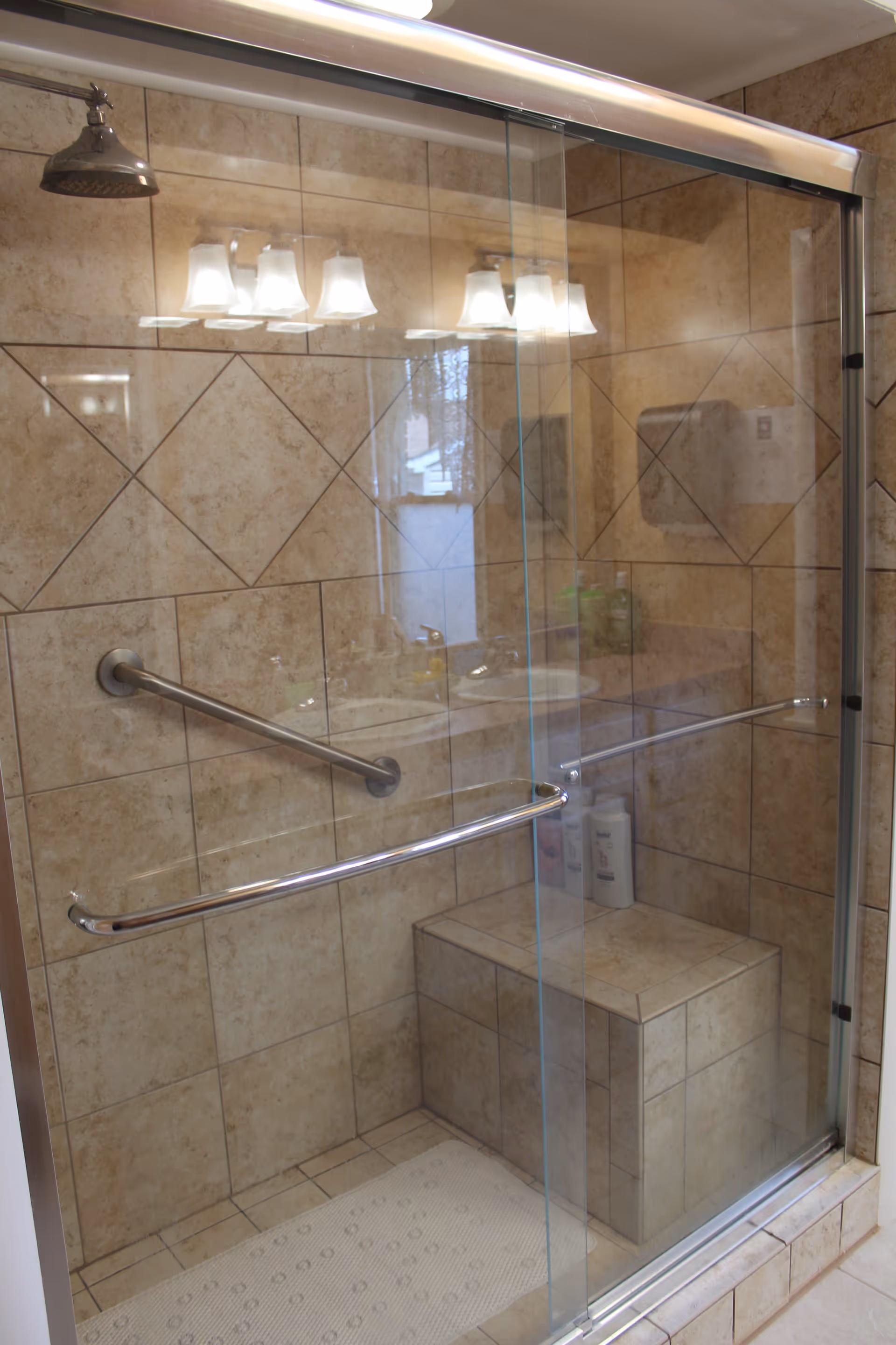 A tiled shower area with a built-in bench, glass sliding doors, and two metal grab bars. The shower has a wall-mounted showerhead and a non-slip mat on the floor. The tiles are beige with a diamond pattern on the walls.