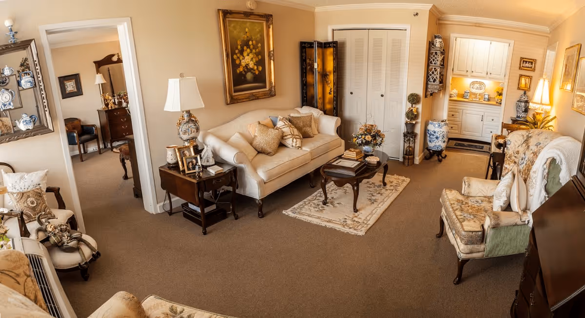 A cozy living room with beige walls and carpet, featuring a cream-colored sofa with decorative pillows, a wooden coffee table on a small rug, and two upholstered armchairs with floral patterns. There is a side table with a lamp and framed photos, a painting of flowers on the wall, and a decorative folding screen near a closet with white louvered doors. In the background, a small kitchenette area with white cabinets and warm lighting is visible.