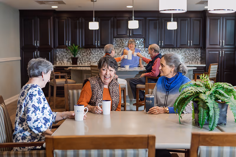 Three elderly women sitting around a table in a kitchen or dining area, smiling and holding coffee mugs. In the background, two elderly people are sitting at a kitchen island, raising their mugs in a toast. The room has dark wooden cabinets and a mosaic tile backsplash.