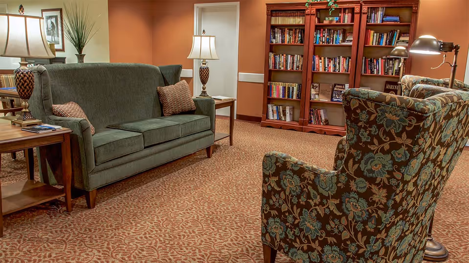 Cozy lounge area with a green sofa, patterned armchairs, side tables and lamps, and a bookshelf against a warm-colored wall.