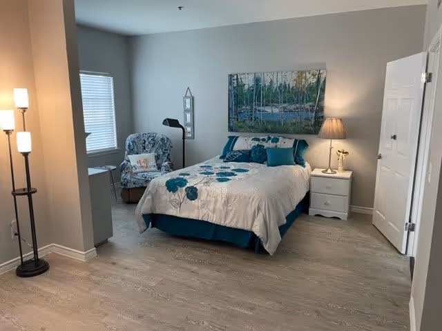 A cozy bedroom with a bed covered in a white and teal floral comforter, a white nightstand with a lamp and a small decorative item, a cushioned armchair with a patterned cover and a pillow, a floor lamp with three lights, and a large window with blinds. The walls are painted light gray and there is a large landscape painting hanging above the bed.