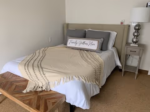 A neatly made bed with a beige throw blanket and multiple pillows, including one with the text 'Family Gathers Here'. Next to the bed is a small nightstand with a silver lamp and a remote control on top. The room has beige carpeting and plain light-colored walls.