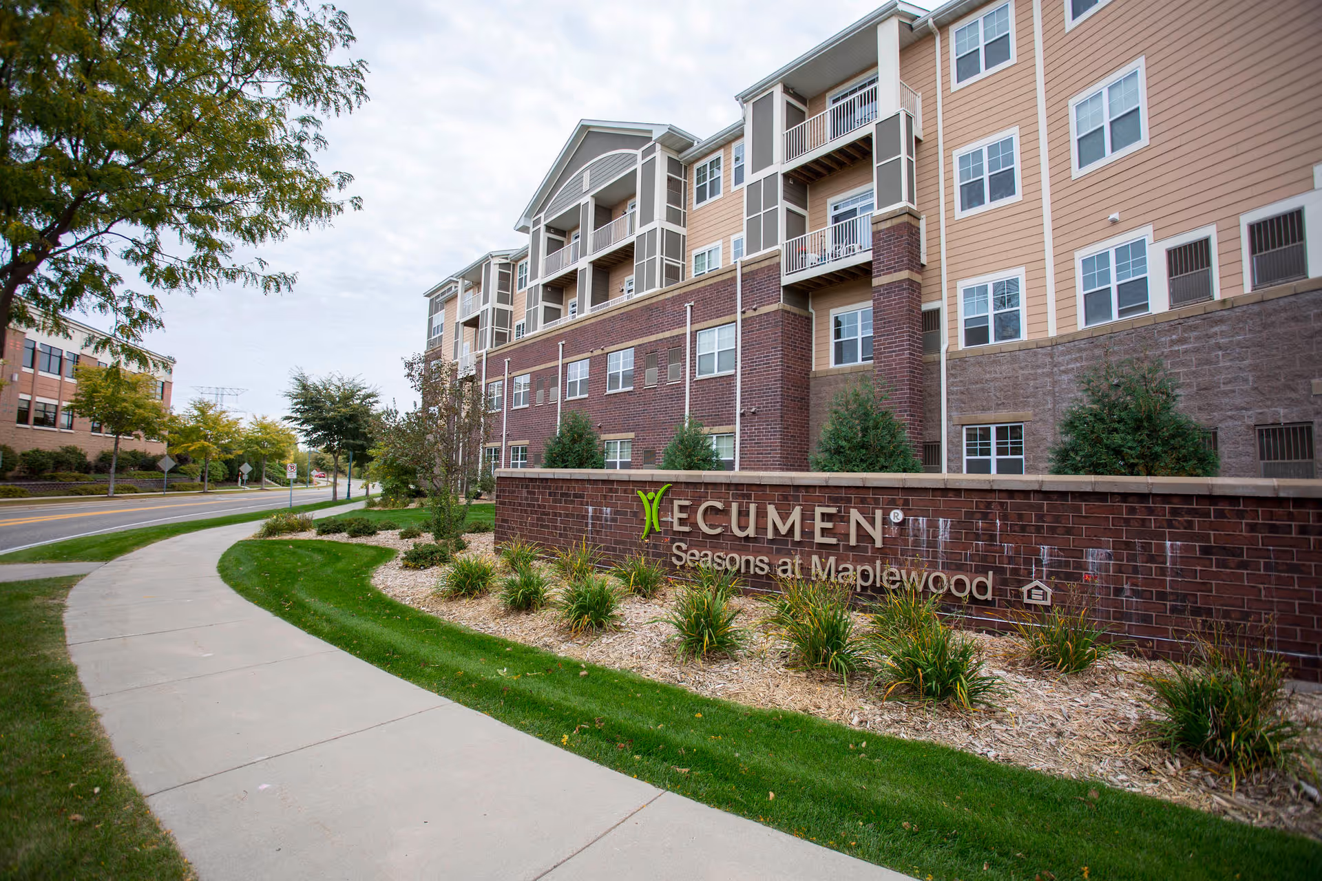 Exterior front of the Ecumen Seasons at Maplewood senior living building with a brick sign, landscaping, and a curved sidewalk.