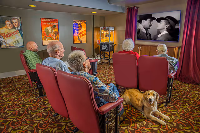 A group of elderly residents sit in red theater chairs watching a black-and-white movie on a screen in a cozy media room while a golden retriever lies on the patterned carpet.