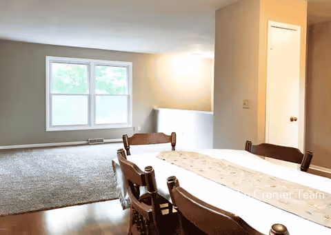 Interior view of a senior living facility showing a dining area with a wooden table and chairs in the foreground and a carpeted living room area with a large window in the background. The walls are painted beige, and there is a door and a small wall partition visible.