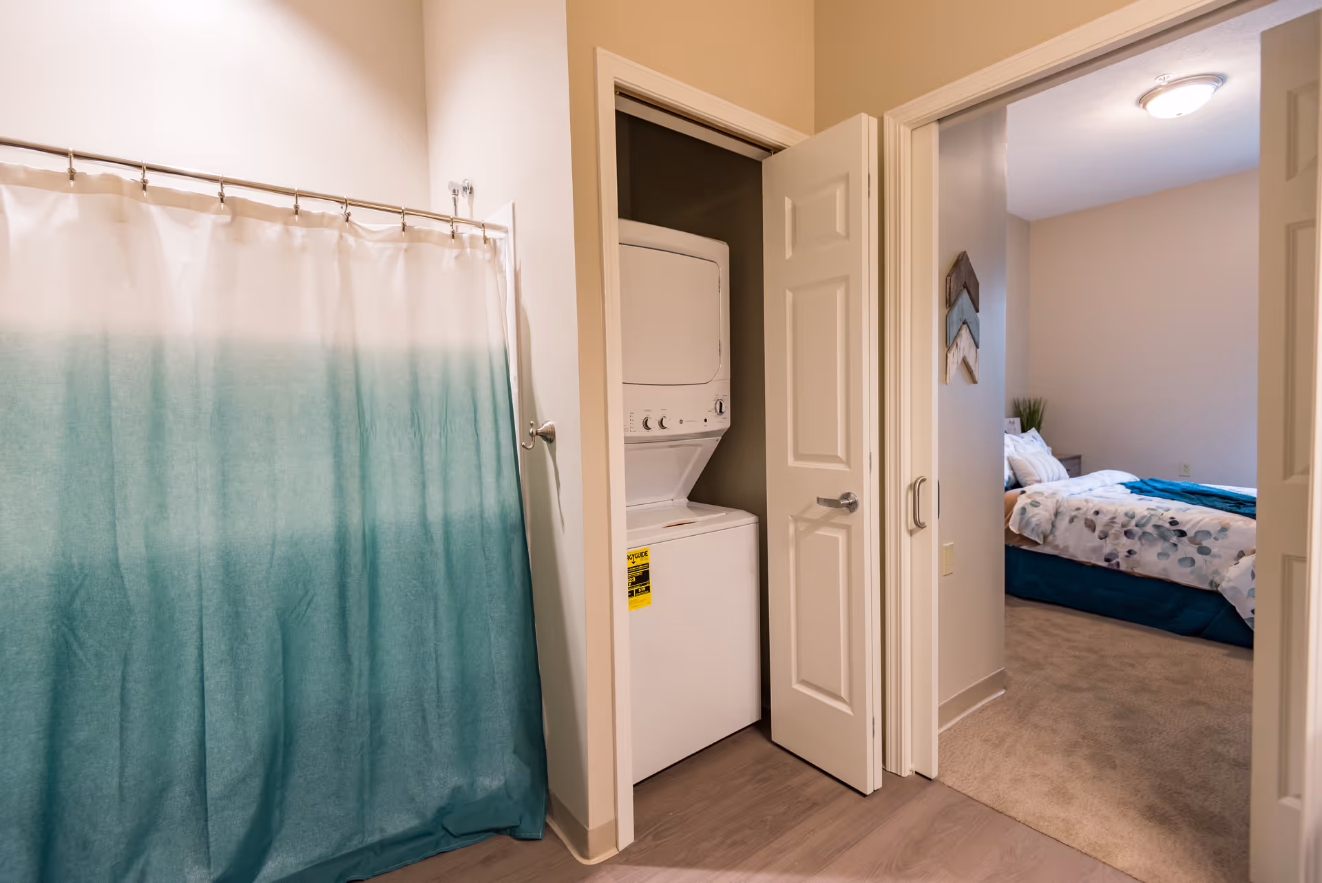 View of a bathroom with a teal and white gradient shower curtain on the left, a small laundry closet with a stacked washer and dryer in the center, and an open door leading to a bedroom with a bed covered in white and blue bedding on the right.