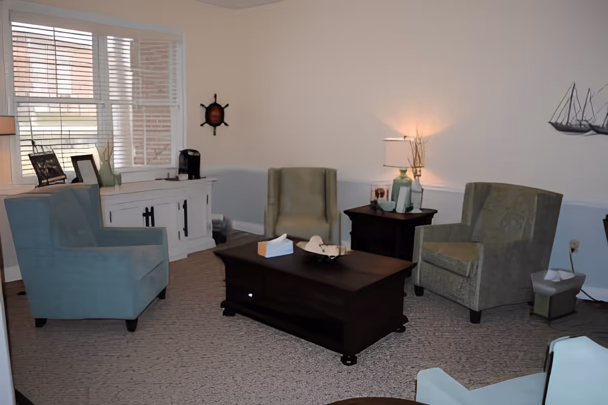 A cozy living room area with four armchairs arranged around a dark wooden coffee table. There is a white cabinet under a window with blinds, a side table with a lamp and decorative items, and wall decorations including a ship's wheel and a metal sailboat sculpture.